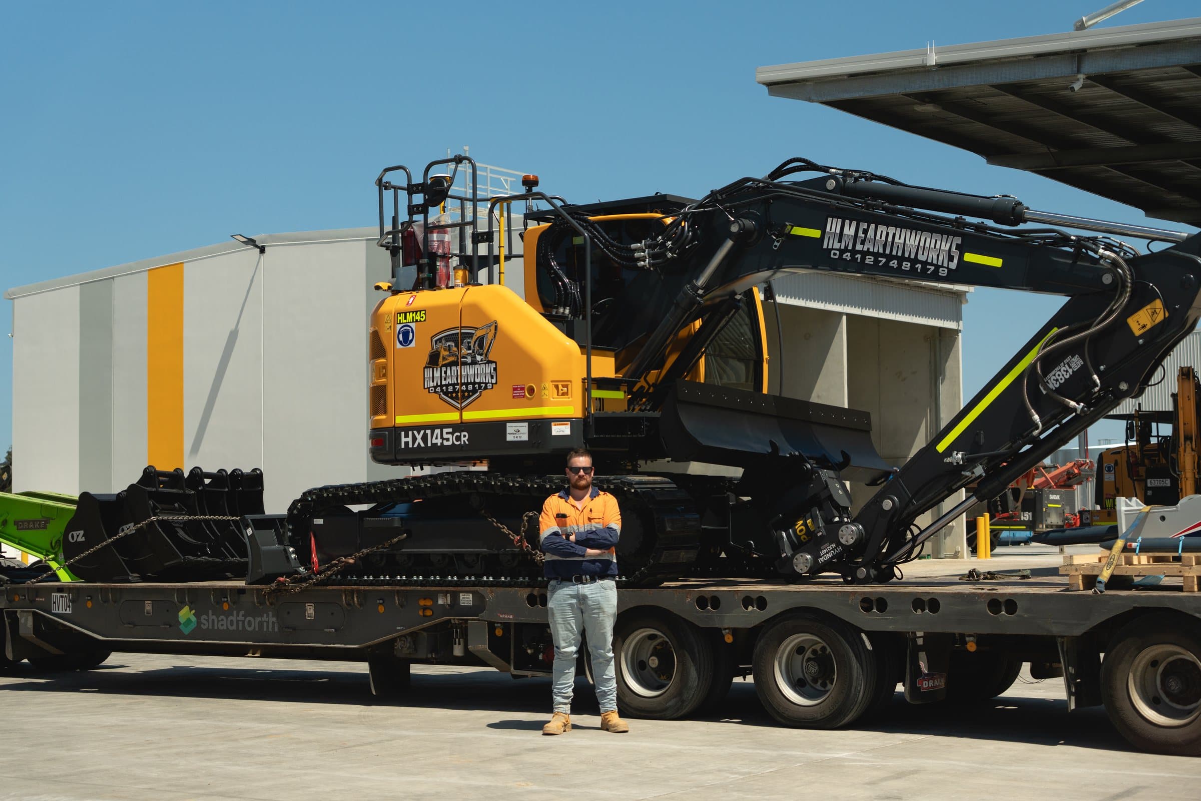 HLM Earthworks crew member in a high-visibility shirt standing in front of a Hyundai HX145CR excavator loaded on a float.
