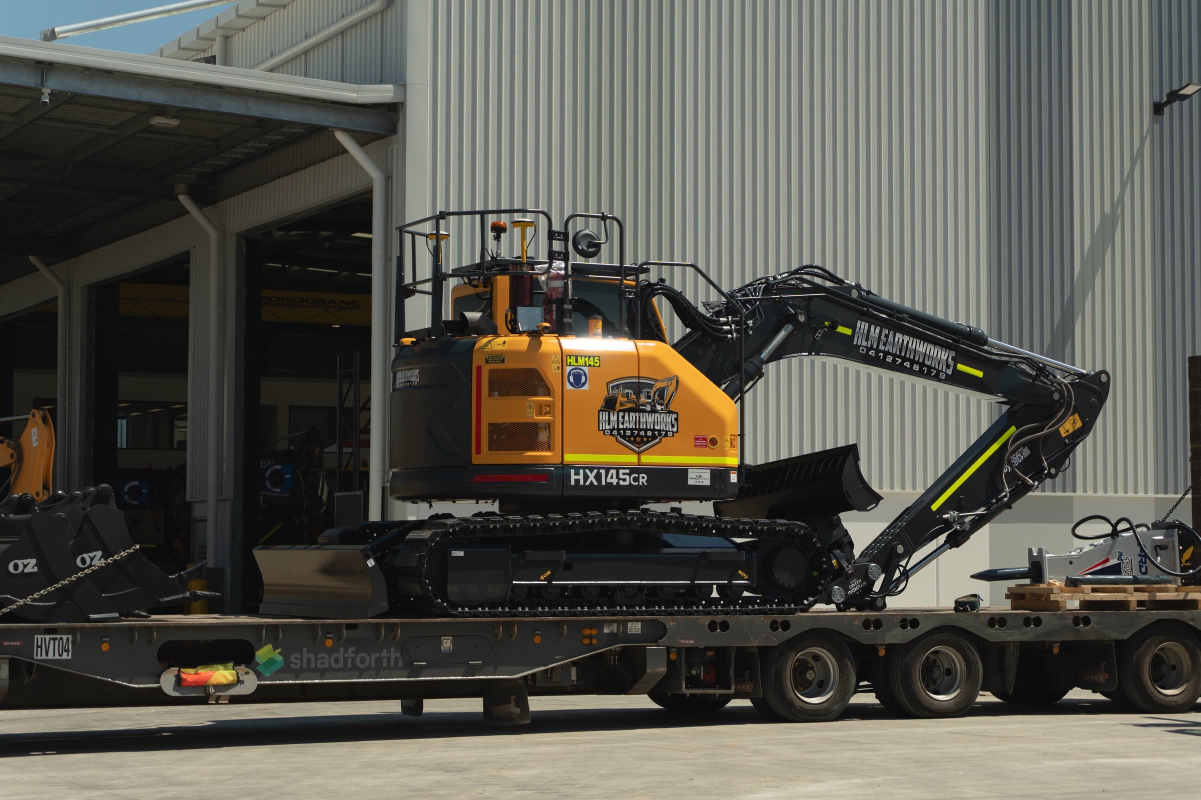 Hyundai HX145CR excavator in HLM Earthworks colours chained to a low-loader outside the workshop.