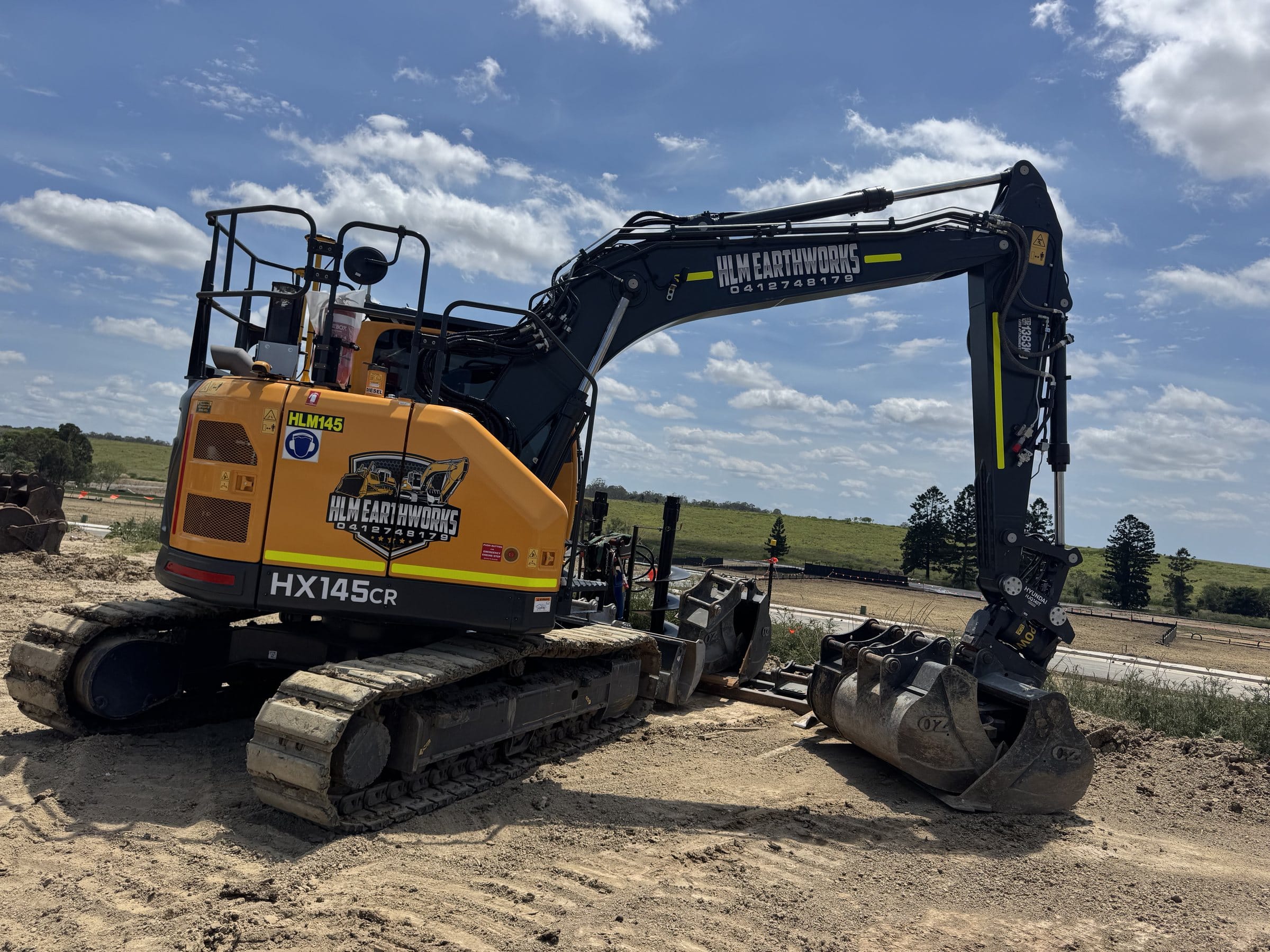 Hyundai HX145CR excavator in yellow and black HLM Earthworks livery, parked on a sandy civil site with green hills and a bright blue sky.