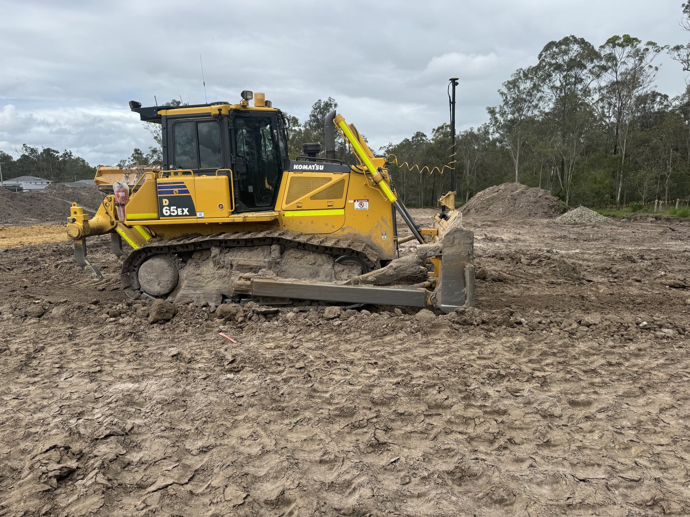 Komatsu D65EX dozer fitted with a GPS machine-control mast, trimming a clay pad beside stockpiles and trees.